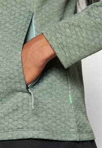 Raižots za�ļais džemperis ar pusi rāvējslēdzēja, satur kabatu, ribotas piedurknes un smalku rakstu uz virsmas, kas piešķir papildus dimensiju.