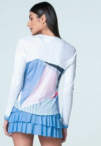 Frau trägt ein langärmliges weißes Oberteil mit geometrischen blauen, rosa und schwarzen Mustern, kombiniert mit einem blauen, gestuften Volantrock, der von hinten zu sehen ist.