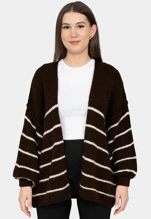 Jeune femme portant un cardigan en maille marron oversize avec des rayures horizontales blanches, par-dessus une chemise blanche et un pantalon noir.