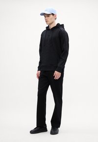 Μαύρη ζακέτα με υφή, μπροστινές τσέπες και κορδόνια. Συνδυασμένη με μαύρο παντελόνι και μαύρα αθλητικά παπούτσια. Ανοιχτό μπλε καπέλο με λογότυπο.