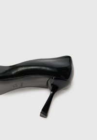 Scarpa nera con tacco alto, materiale in pelle verniciata liscia, punta affilata, tacco stiletto sottile con punta in gomma testurizzata, design minimale.