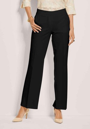 Vrouw draagt zwarte rechte broek met een crèmekleurige gestructureerde blouse, beige hoge hakken, een armband en een ring, staande tegen een lichte achtergrond.