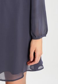 Ruka odpočívající vedle okraje průhledné dlouhé modré šatů s elastickým manžetovým lemem a poloprůhledným látkovým detailem.