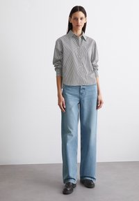 Gestreiftes Hemd in Hellgrau und Weiß, kombiniert mit weitem hellblauem Jeans und schwarzen Plateauschuhe, vor einem neutralen Hintergrund.