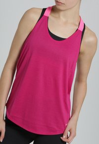 Femme portant un débardeur de sport rose sans manches par-dessus un soutien-gorge de sport noir, avec des leggings noirs, posant devant un fond uni.