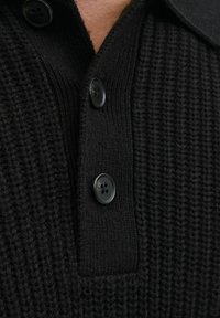Polo de punto negro con una tela texturizada, que presenta un collar y dos botones negros en la tapeta. Primer plano del material.