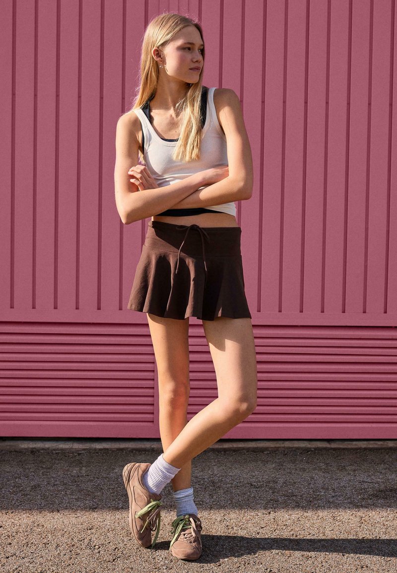 Jeune femme aux longs cheveux blonds, bras croisés, portant un débardeur blanc, une jupe marron et des baskets, devant un mur rose.