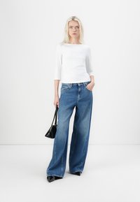 Bredde ben jeans i blå denim med en falmet finish, kombinert med en tettsittende hvit topp med trekvart lange ermer. Modellen holder en liten svart håndveske.