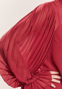 Rød bluse med gennemsigtig, plisseret stof på ærmerne og rynkedetaljer ved håndleddene. Den glatte tekstur supplerer designet.