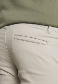 Beige cotton trousers featuring a discreet back pocket, smooth texture, and minimal stitching details, worn with a green sweatshirt.