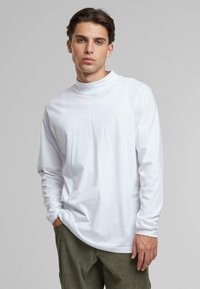 Camiseta blanca de manga larga con un cuello alto, hecha de algodón ligero. Presenta un corte holgado y una textura suave, combinada con pantalones de pana verdes.