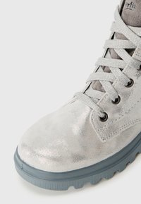 Bottine métallique argentée avec une texture lisse, lacets gris et semelle en caoutchouc grise épaisse. Présente un bout rond et de petites perforations.