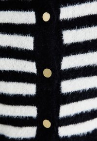 Svartvit randig kofta i mjukt stickat tyg, med runda guldfärgade knappar längs fram. Randernas bredd varierar.