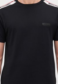 T-shirt in cotone nero con scollo a girocollo, maniche corte e bretelle bianche a contrasto con il testo "MOSCHINO" lungo le spalle.