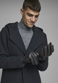 Guantes de cuero negro con una textura suave, usados por una persona con un abrigo oscuro y un cuello de tortuga gris, preparándose para ponérselos.