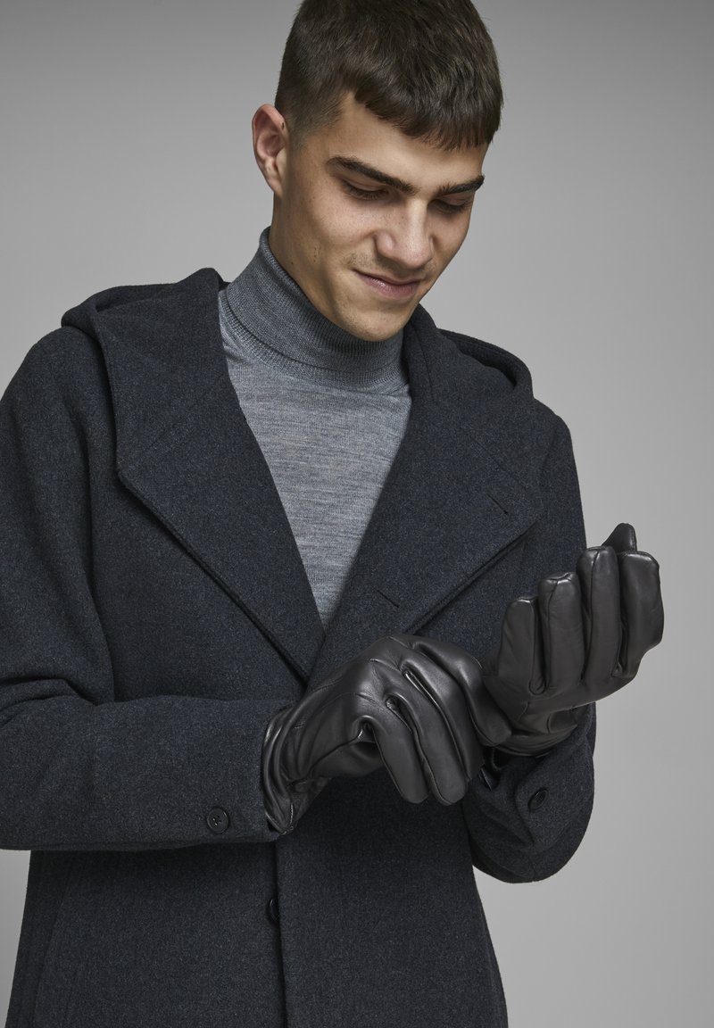 Guantes de cuero negro con una textura suave, usados por una persona con un abrigo oscuro y un cuello de tortuga gris, preparándose para ponérselos.
