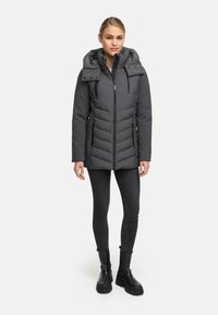 Grijze pufferjack met ritssluiting, chevronpatroon, afneembare capuchon, zijzakken en een glanzende afwerking, gedragen over zwarte jeans en laarzen.