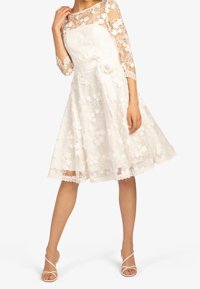 Robe blanche en dentelle avec des manches trois-quarts, broderies florales, un corsage ajusté et un nœud à la taille. Jupe en ligne A avec finition en dentelle au bas.