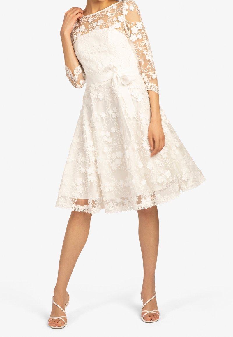 Robe blanche en dentelle avec des manches trois-quarts, broderies florales, un corsage ajusté et un nœud à la taille. Jupe en ligne A avec finition en dentelle au bas.