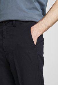 Námořnické modré bavlněné kalhoty s hladkou texturou, předními kapsami a knoflíkovým zapínáním. Viditelné sklady látky v pase.