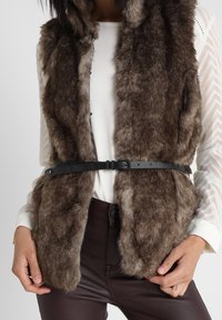 Braune Kunstpelzweste mit weicher, flauschiger Textur, die mit einem schwarzen Gürtel geschlossen wird. Die Weste hat Knöpfe vorne.