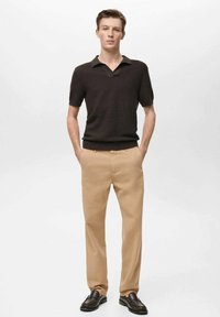 Rjav teksturiran polo majica, bež hlače in temni loaferji. Model pozira z rokami v žepih proti enotnemu ozadju.