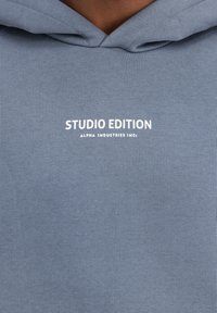 Hoodie grigio in cotone con texture a coste e logo frontale bianco con la scritta: "STUDIO EDITION" e "ALPHA INDUSTRIES INC."