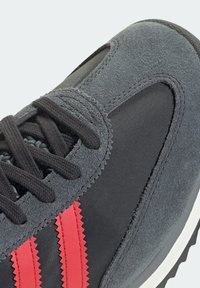 Grå sneakers med en blandning av mocka och textil, svarta snören, röda texturerade ränder och en vit gummisula med en texturerad yttersula.