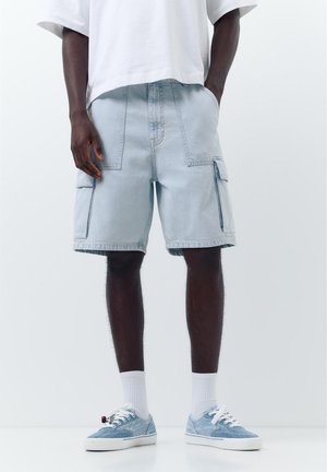 Mand iført lyseblå denim cargo shorts, hvide crew sokker, blå sneakers og en hvid t-shirt, stående med den ene hånd i lommen.