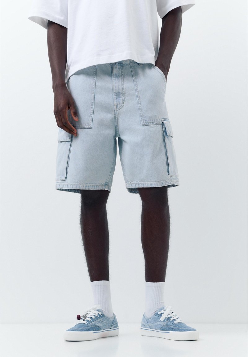 Homme portant un short cargo en jean bleu clair, des chaussettes blanches, des baskets bleues et un t-shirt blanc, debout avec une main dans la poche.