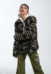Giacca in pile con motivo camouflage verde e beige, chiusura con zip, colletto alto; abbinata a pantaloni verde oliva con ricami decorativi.