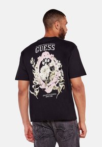 Czarny bawełniany t-shirt z kwiatowym wzorem nadrukowanym na białym i różowym tle na plecach. Zawiera nazwę marki "Guess" oraz napis "Quality Clothing".