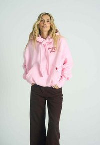 Růžová mikina s kapucí, klokaní kapsou a šňůrkami, s červeně vyšitým textem. Kombinována s hnědými rozevlátými kalhotami.