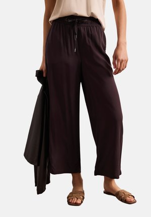 Donna che indossa pantaloni neri corti e larghi con coulisse in vita, top beige, sandali intrecciati color cuoio, che tiene una giacca nera nella mano sinistra.