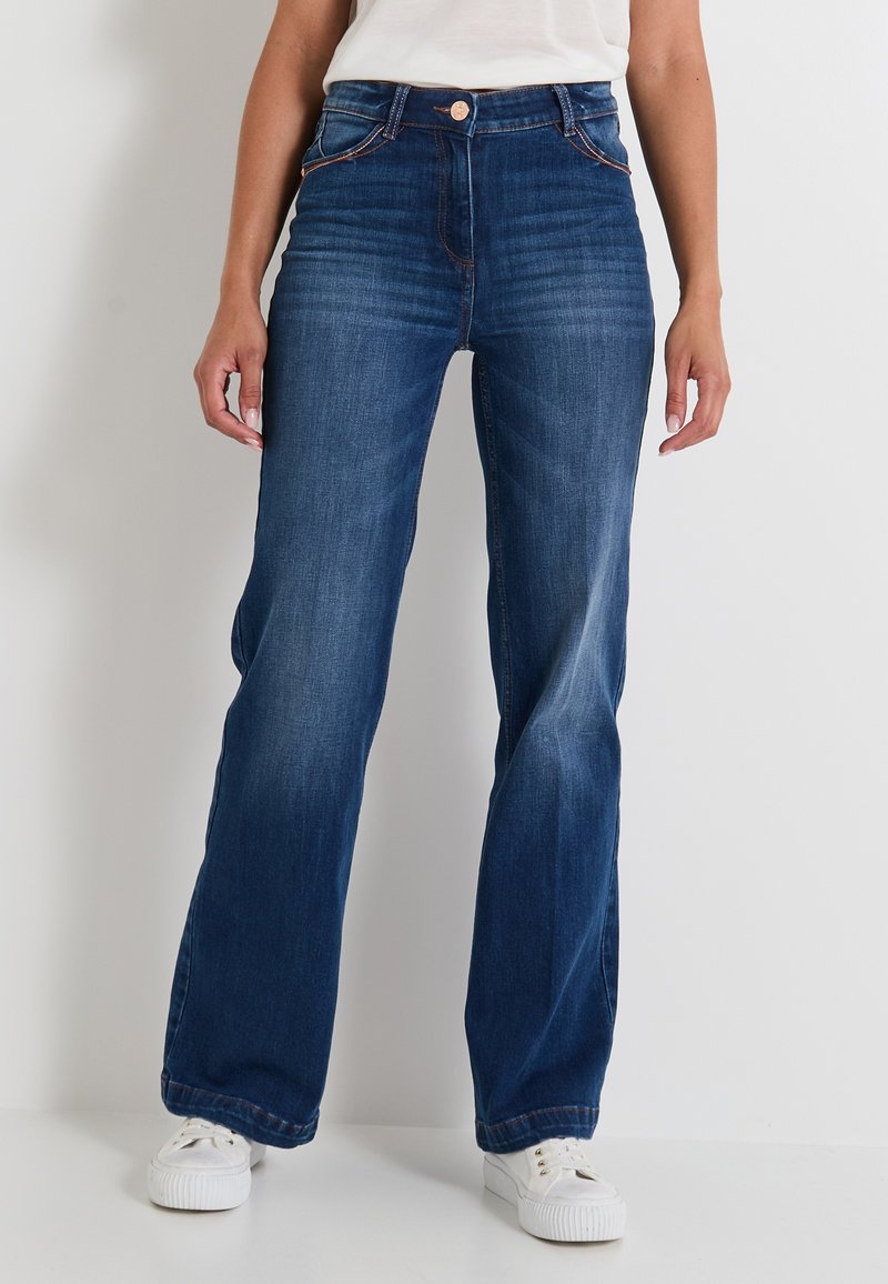 Jean en denim bleu foncé, au design taille large, avec des poches avant et des coutures visibles. Associés à des chaussures blanches pour le contraste.