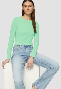 Haut vert clair à manches longues avec une coupe ajustée, associé à un jean bleu clair et des chaussures blanches. Modèle assis sur un bloc blanc.