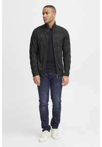Mørk grå zip-up genser med høy krage, kombinert med en svart skjorte, blå jeans og hvite sneakers. Genseren har et teksturert design.