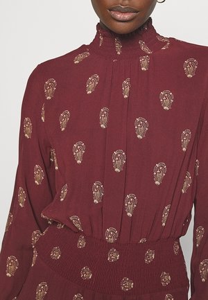 Kvinde iført en mørkerød kjole med lange ærmer med en høj, flæset krave og gyldne paisley-mønstre, der sidder tæt til i taljen med smock-detaljer.