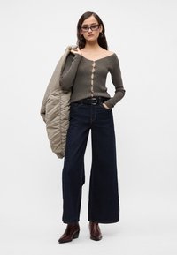 Γυναίκα φορώντ�ας σκούρες φαρδιές παντελόνες, γκρι πουλόβερ με κουμπιά και ανοιχτούς ώμους, γυαλιά ηλίου, κρατώντας ένα μπεζ μπουφάν πάνω από τον ώμο, και καφέ μποτάκια μέχρι τον αστράγαλο.