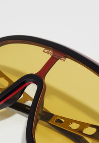 Zonnebril met een zwart en rood frame, gele lenzen en een slank ontwerp. Heeft uitsparingen op de armen en een zichtbaar merklogo op de lens.