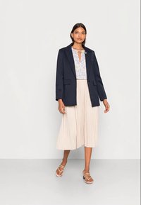 Blazer de tecido macio azul-marinho com lapelas recortadas, combinado com uma blusa padrão azul claro e uma saia midi plissada em creme fluida.