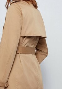 Beżowy trencz z szerokim kołnierzem i paskiem w talii, wykonany z gładkiej, teksturowanej tkaniny, z plisowanym detalem z tyłu.
