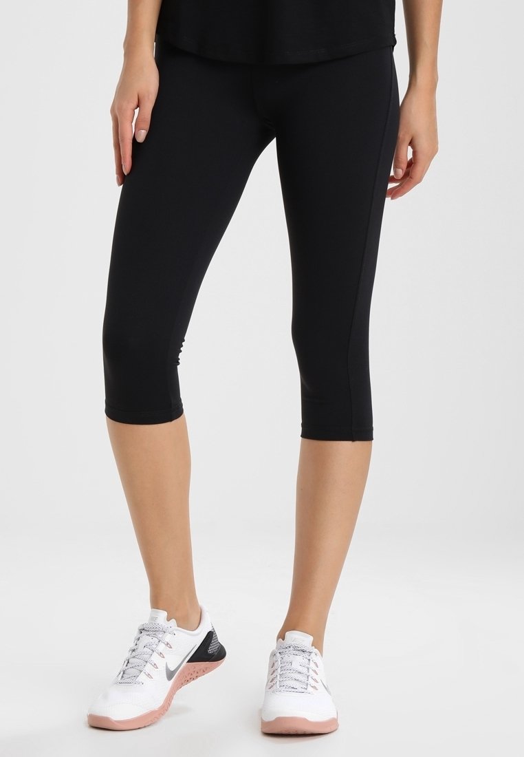 Femme portant des leggings noirs courts et des chaussures de sport blanches à semelles roses, debout devant un fond blanc.