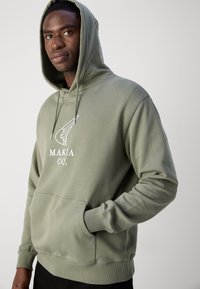 Sweat à capuche vert olive avec cordon de serrage, poches avant et logo blanc comportant des ailes stylisées. Texture douce et coupe décontractée.