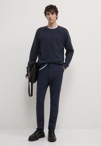 Felpa navy, pantaloni scuri e scarpe nere. Texture morbida, girocollo e design minimal. Il modello tiene una borsa nera.