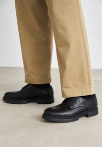 Chaussures en cuir noir à texture lisse, bout arrondi et épaisse semelle en caoutchouc. Associées à un pantalon kaki, avec peu de détails visibles.