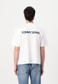 Hvid bomulds T-shirt med "TOMMY JEANS" logo i marineblå på ryggen. Skåret med en afslappet pasform og korte ærmer, parret med blå jeans.