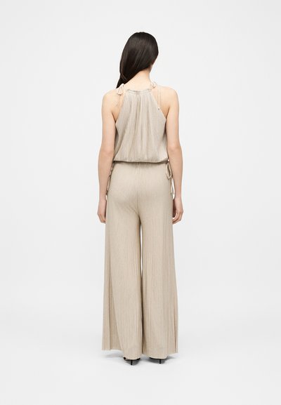 Femme aux longs cheveux foncés, portant un haut beige sans manches à col halter et un pantalon à jambes larges, debout de dos sur un fond uni.