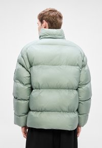 Πράσινο μπουφάν τύπου puffer με ψηλό γιακά και σχέδιο καπιτονέ, με παχιά επένδυση και λεία υφή. Η πίσω όψη αποκαλύπτει μια άνετη εφαρμογή.