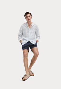 Camisa de manga larga a rayas blancas y azules, pantalones cortos azul marino y sandalias beige con hebillas, de pie contra un fondo blanco.